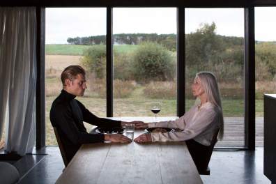 Kunstverk som viser en ung mann og en eldre kvinne sittende ved et bord. Natur, trær og busker i bakgrunnen.