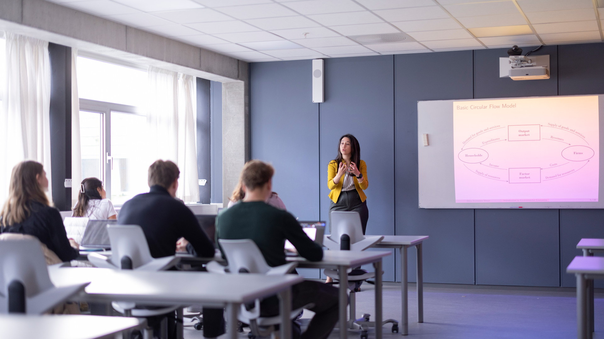 Foreleser ved tavle underviser studenter som sitter ved pultene sine