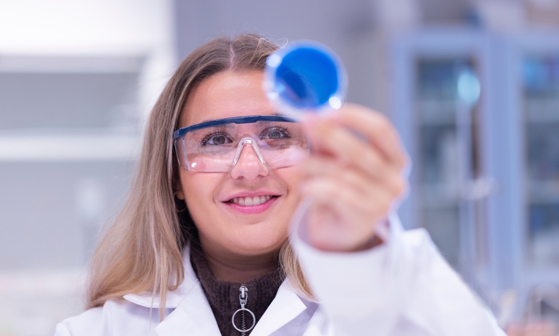 Kvinnelig student iført lab frakk og vernebriller som ser på en beholder med noe blått i