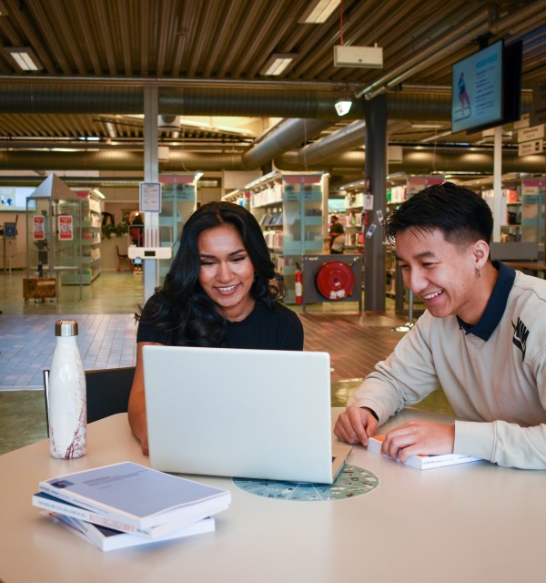 To studenter sitter sammen foran en laptop ved et skrivebord. Bibliotek i bakgrunnen.