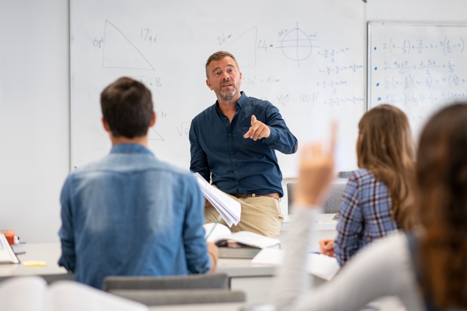 En mannlig lærer står foran i et klasserom med matematiske regneoperasjoner på tavla bak seg