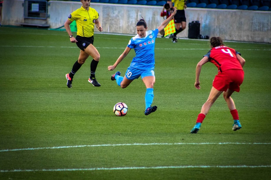 To kvinnelige fotballspillere i kampsituasjon på en grønn fotballbane. Spilleren i blå drakt skal til å skyte, mens spilleren i rød drakt står med ryggen til kamera og forsvarer. I bakgrunnen ses en mannlig dommer i gult.
