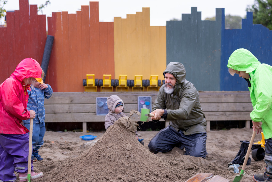 Voksen mann med skjegg og regnjakke leker i sandkassen med tre barn