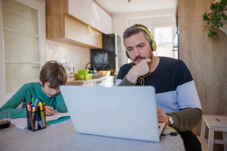 Mann sitter med øretelefoner og datamaskin. Ved siden av sitter et barn og tegner. 