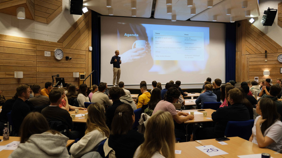 Mann holder foredrag. Studenter sitter rundt flere bord.